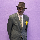 Elder black man in a gray suit, tie, and hat standing in front of a purple wall